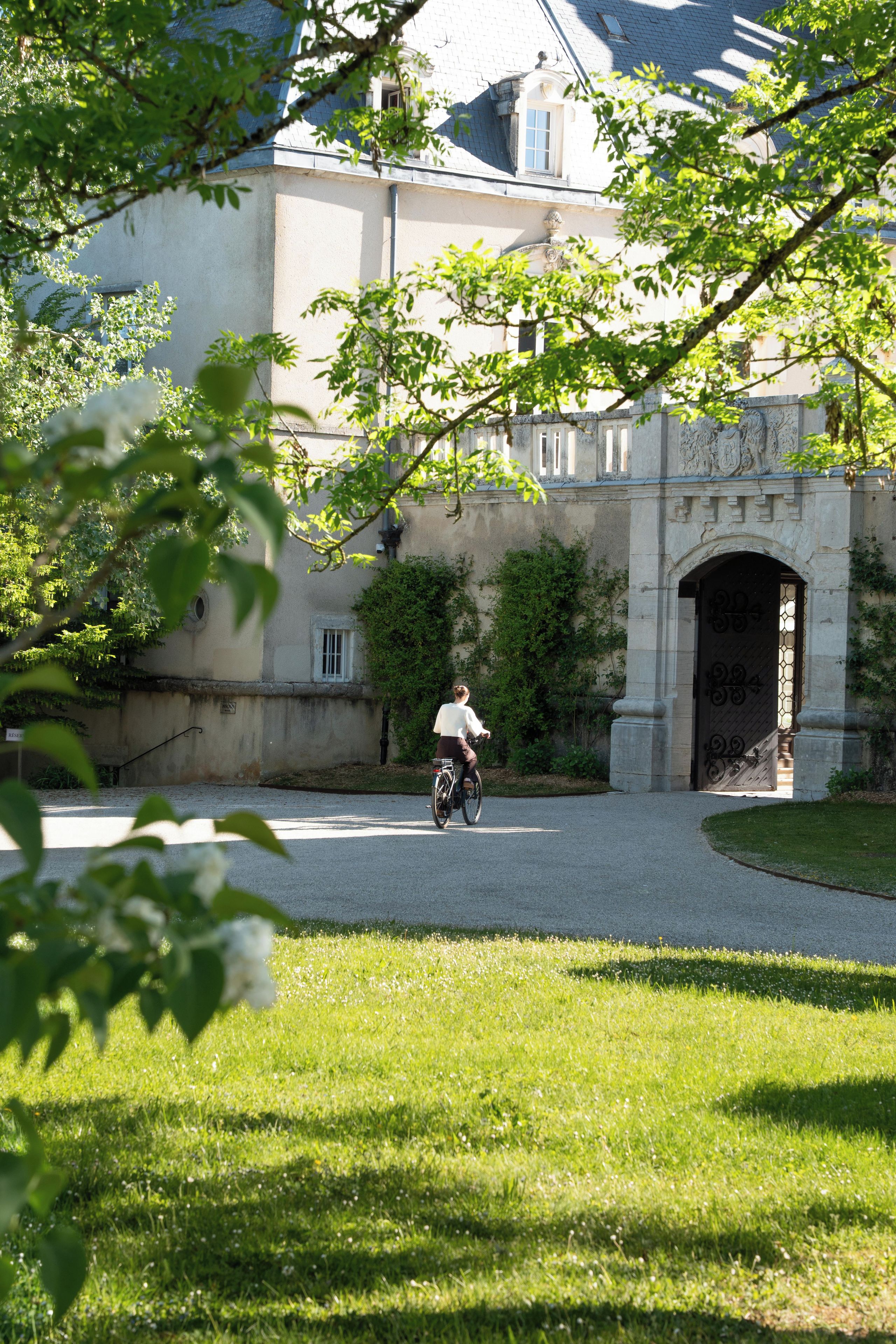 A person riding a bicycle on a paved path in front of a building with a large arched entrance and a stone wall.