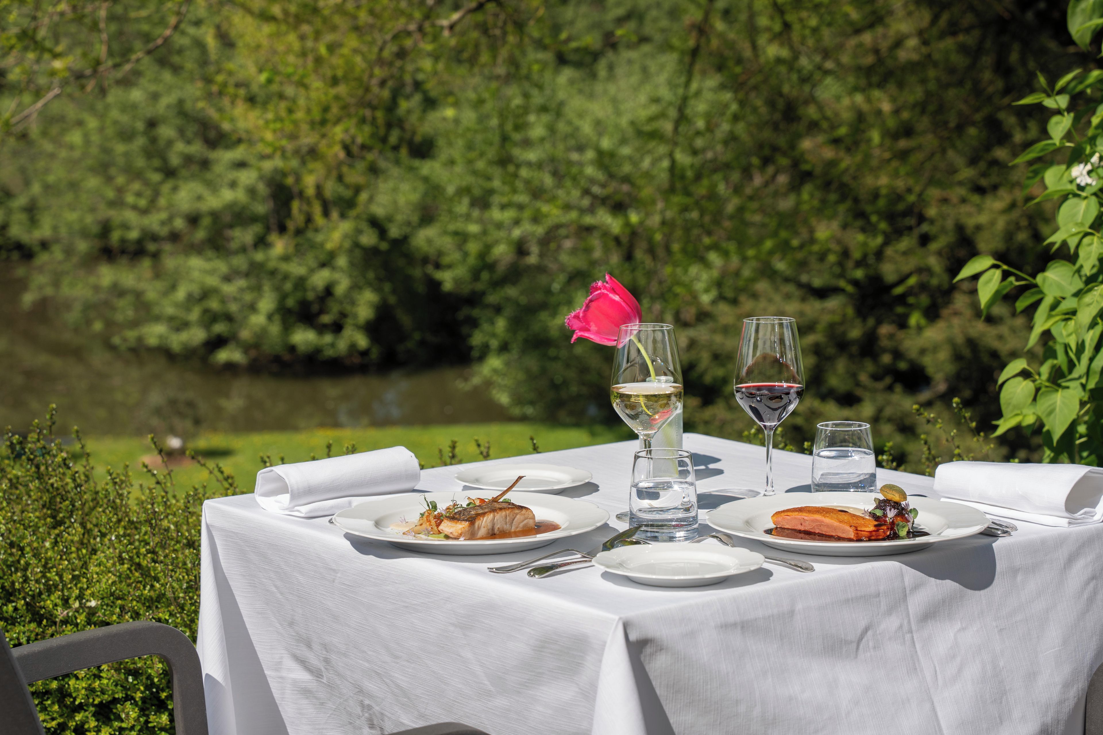 Een buitentafeltje met borden eten, glazen wijn en een roze bloem.