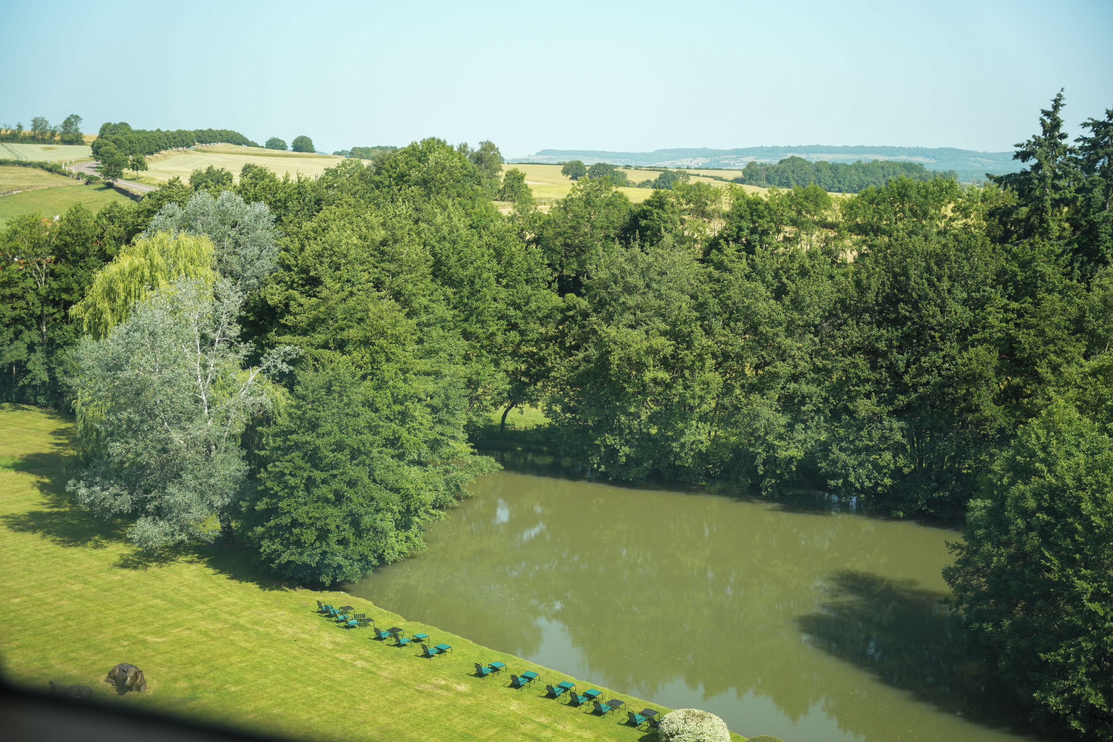 Une vue sur la rivière, avec une zone herbeuse et des arbres, une étendue d'eau calme, et une rangée de chaises le long du bord de l'eau.