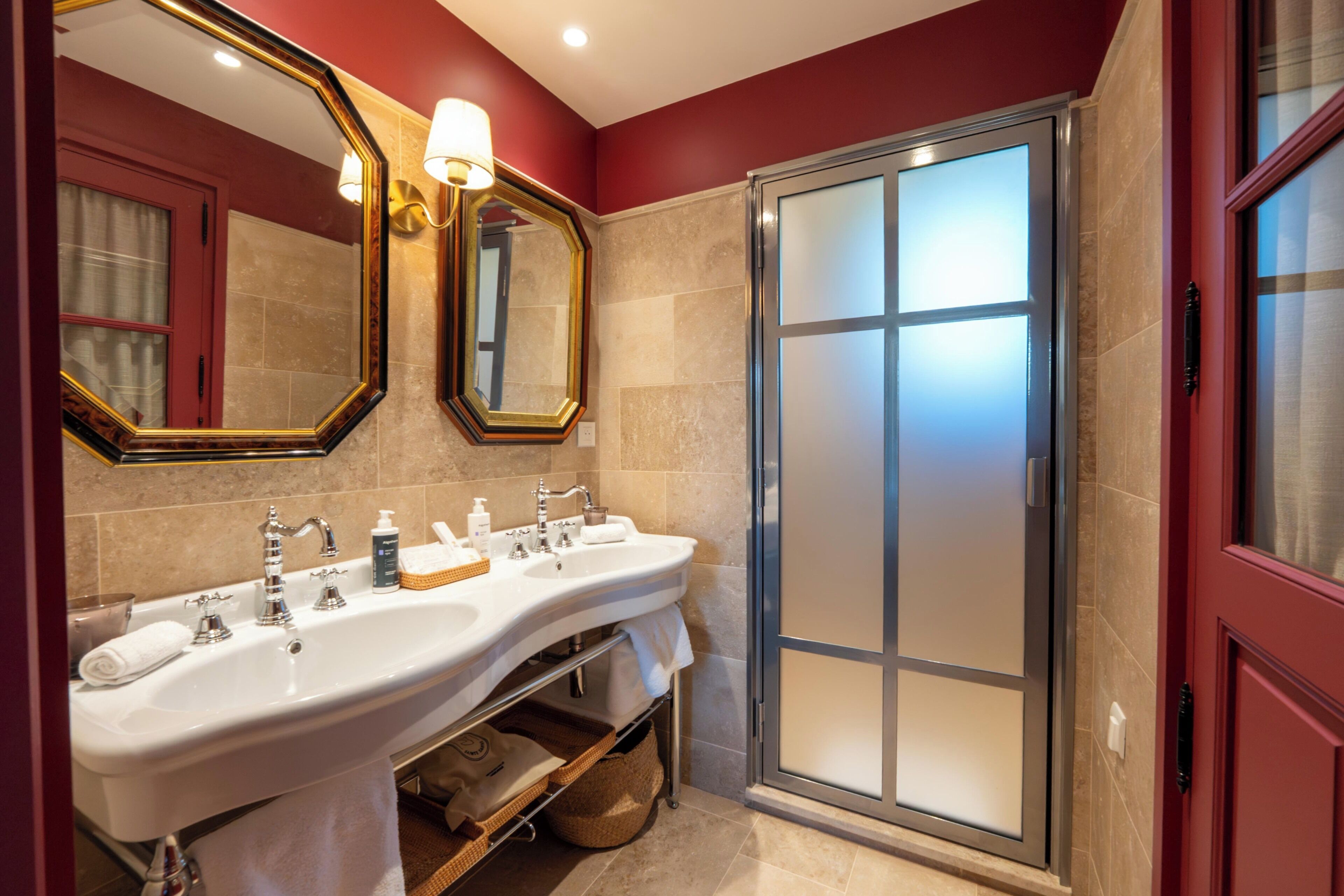Une salle de bain avec un double lavabo, un grand miroir, une douche avec des portes en verre dépoli et un mur d’accent rouge.