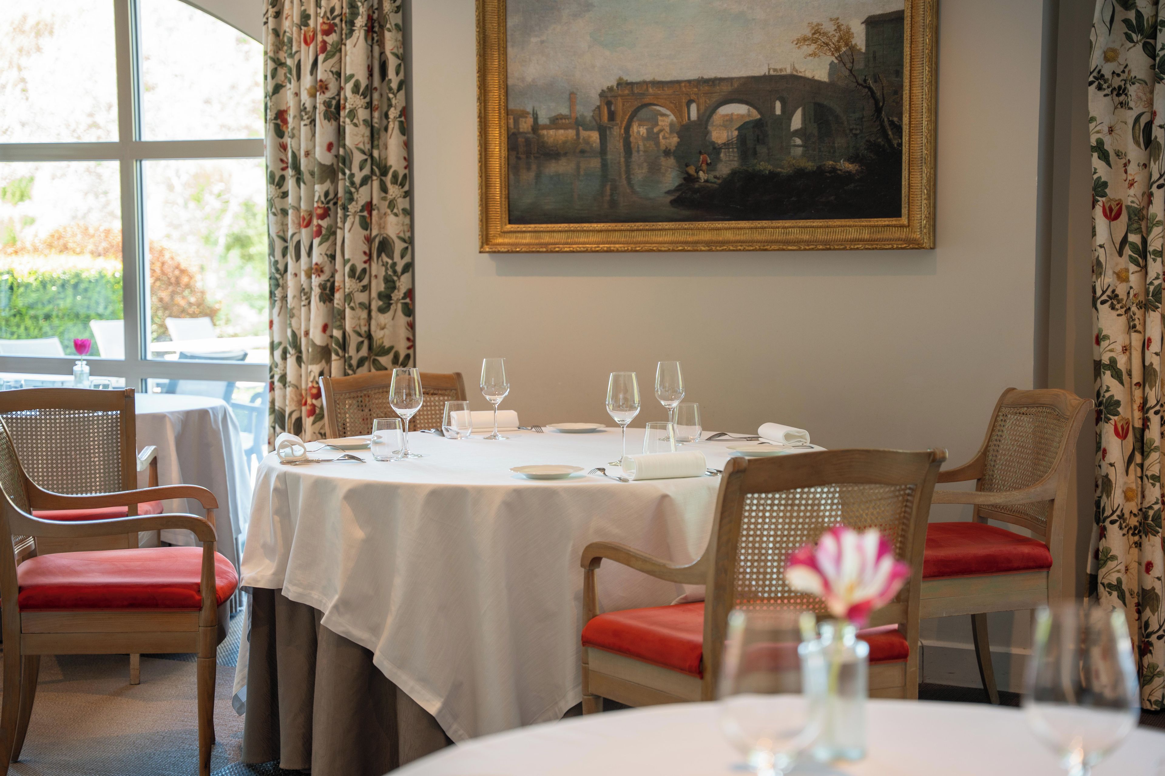 A dining area with a table set for four, chairs with red cushions, and a painting on the wall.