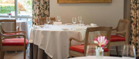 A dining area with a table set for four, chairs with red cushions, and a painting on the wall.