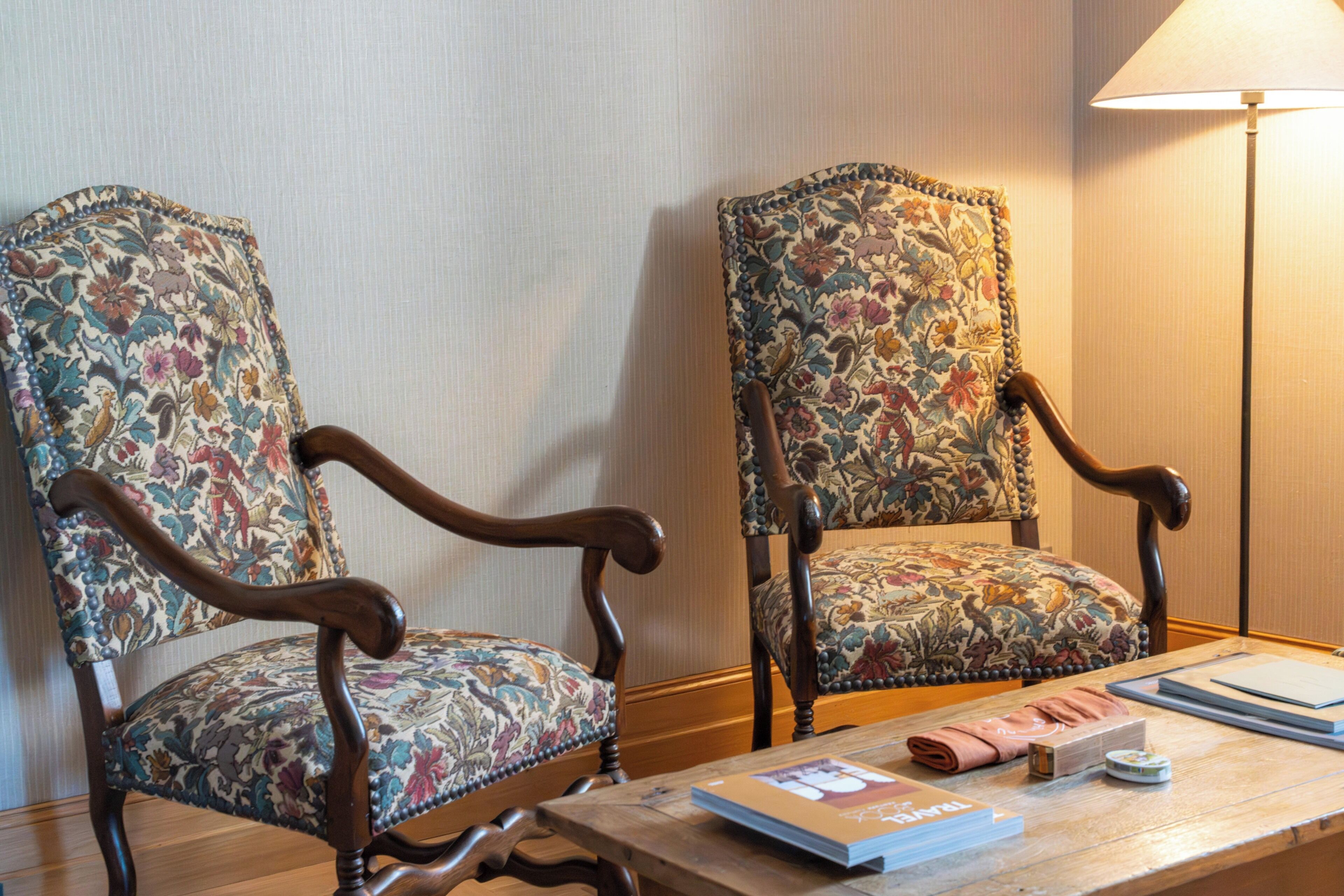 Two upholstered chairs with floral patterns, a wooden table with a book and a rolled-up towel, and a floor lamp.