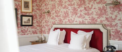 Una cama bien tendida con sábanas blancas y un cabecero rojo, una mesita de noche con una lámpara y un teléfono, y una pared con papel tapiz floral y cuadros enmarcados.