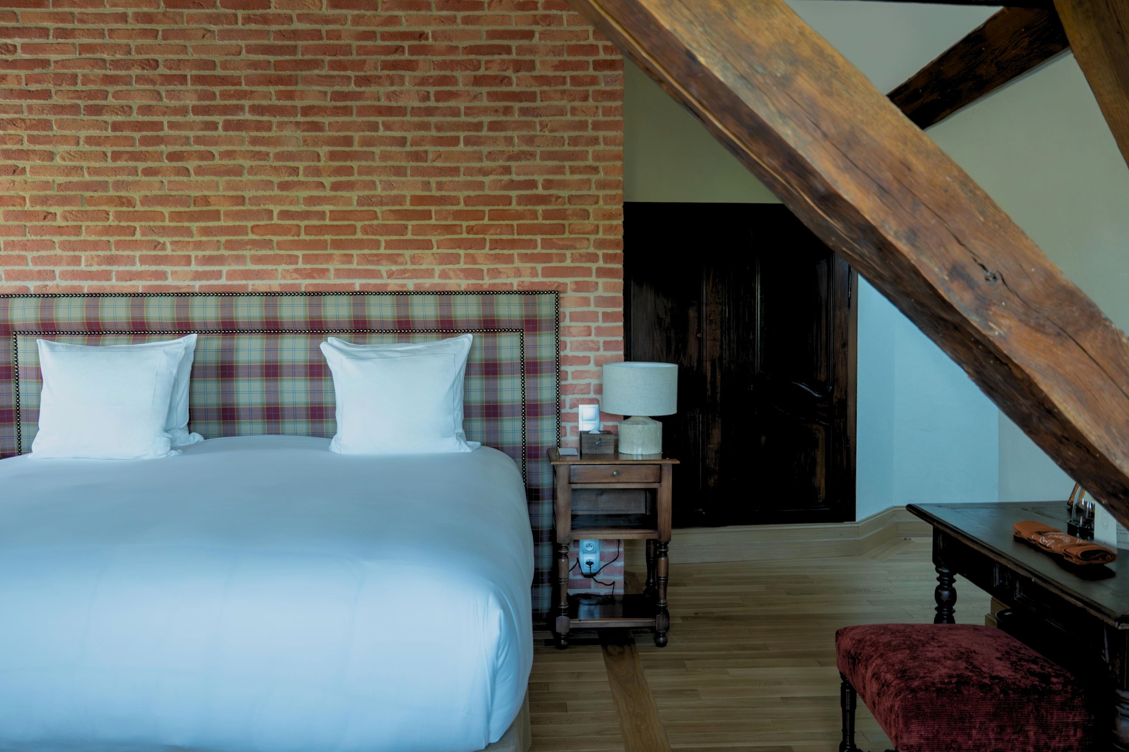 Ein Schlafzimmer mit Backsteinwand, einem Bett mit weißen Leinen, einem Karomuster-Kopfende, einem Nachttisch mit Lampe und einer Holzbank.