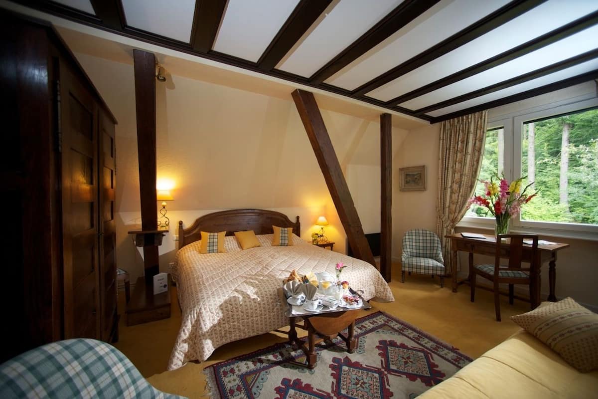 Ein Schlafzimmer mit Bett, Nachttisch, Lampe, Stuhl und einem Fenster mit Blick ins Grüne.