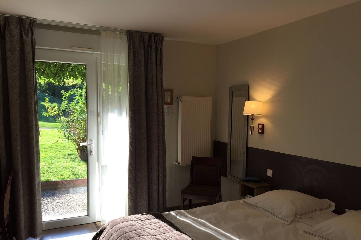 Ein Hotelzimmer mit Bett, Stuhl, Schreibtisch, Lampe und Blick auf einen Garten durch die Tür.