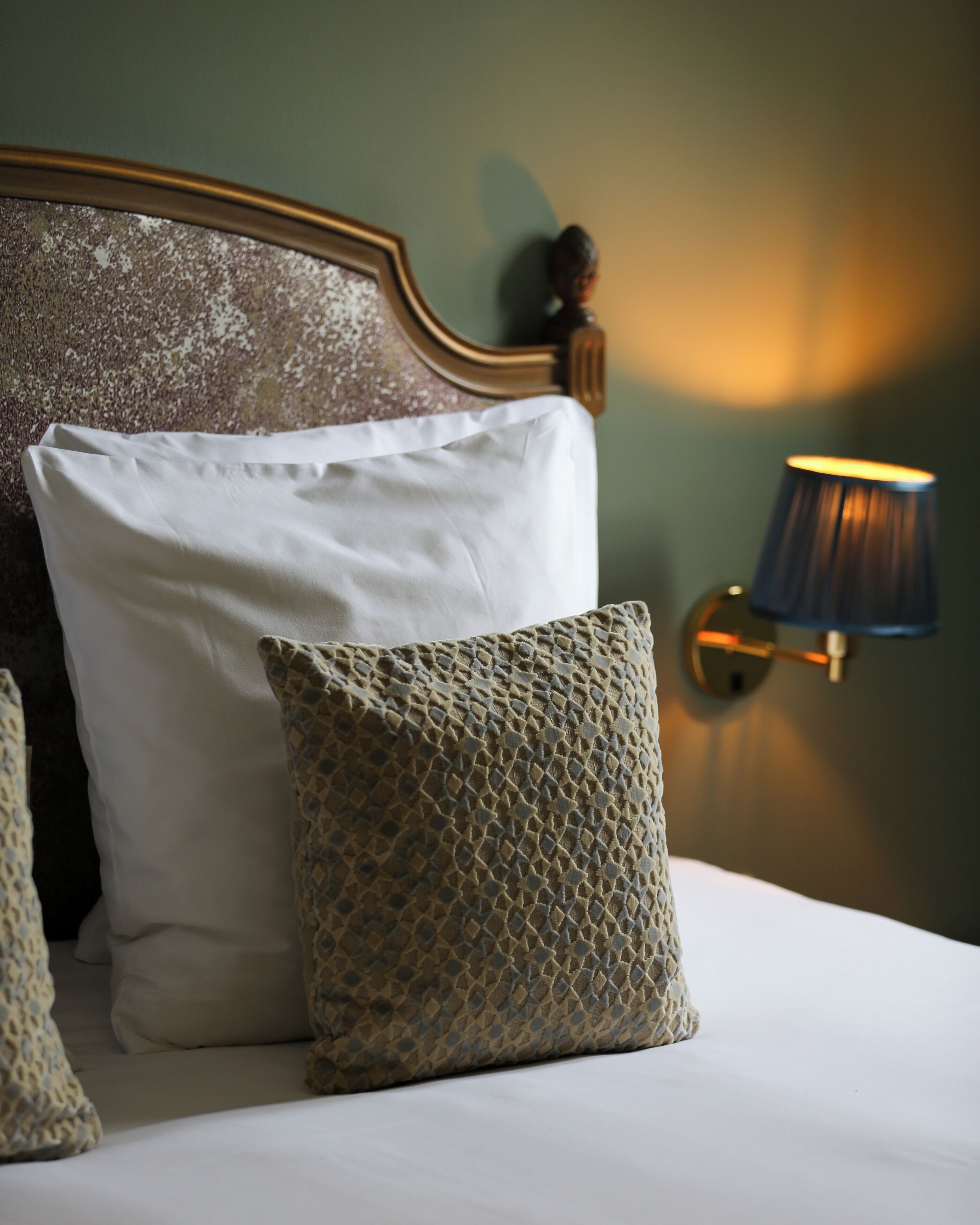 Een bed met witte en gemêleerde kussens, een houten hoofdbord en een wandlamp.