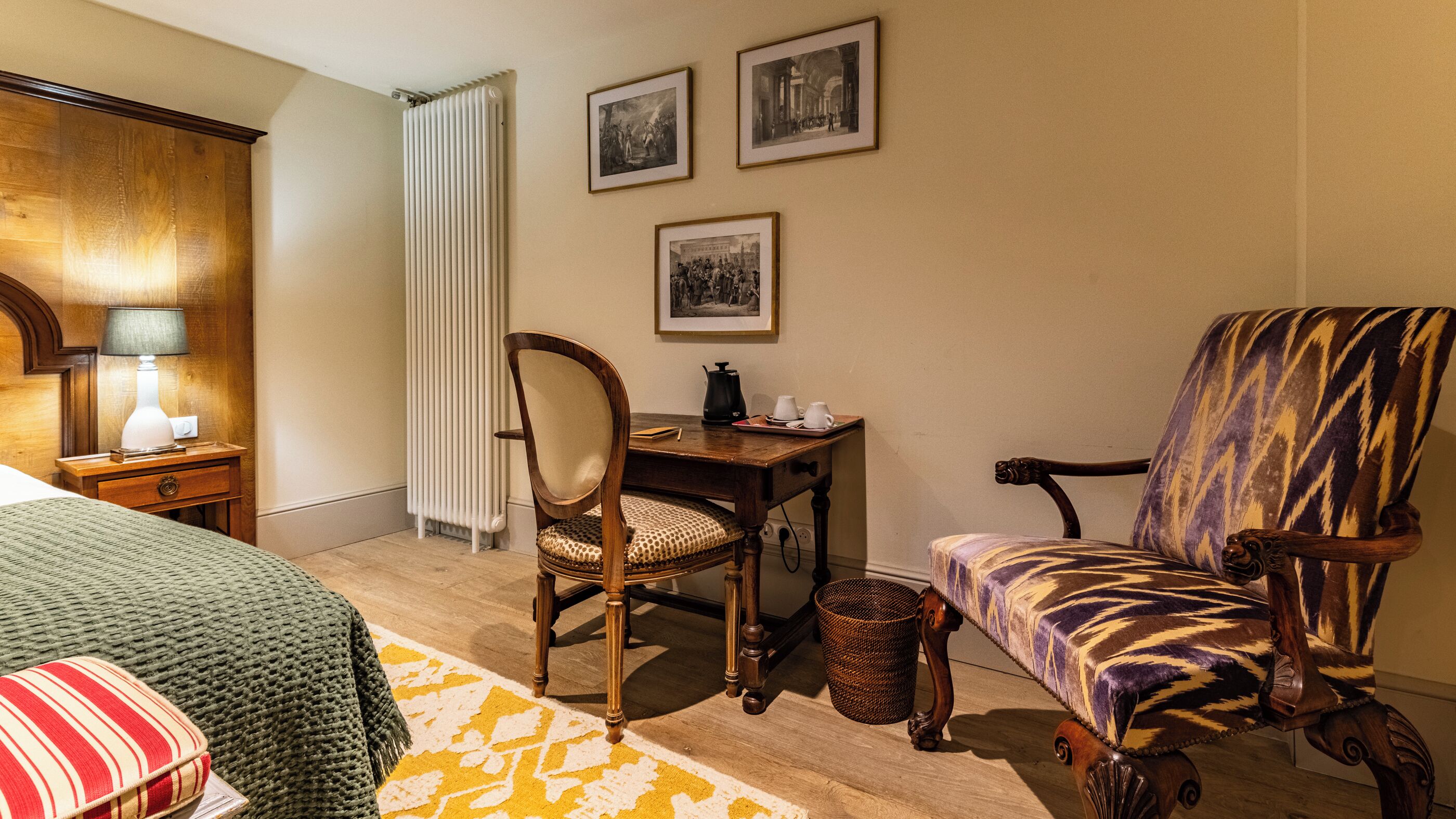 Een hotelkamer met een bed, een bureau met stoel, een radiator en ingelijste foto's aan de muur.