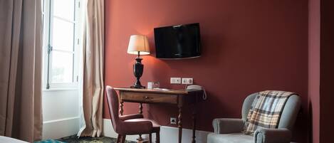A hotel room with a bed, a desk with a lamp, a chair, a television, and a window with curtains.