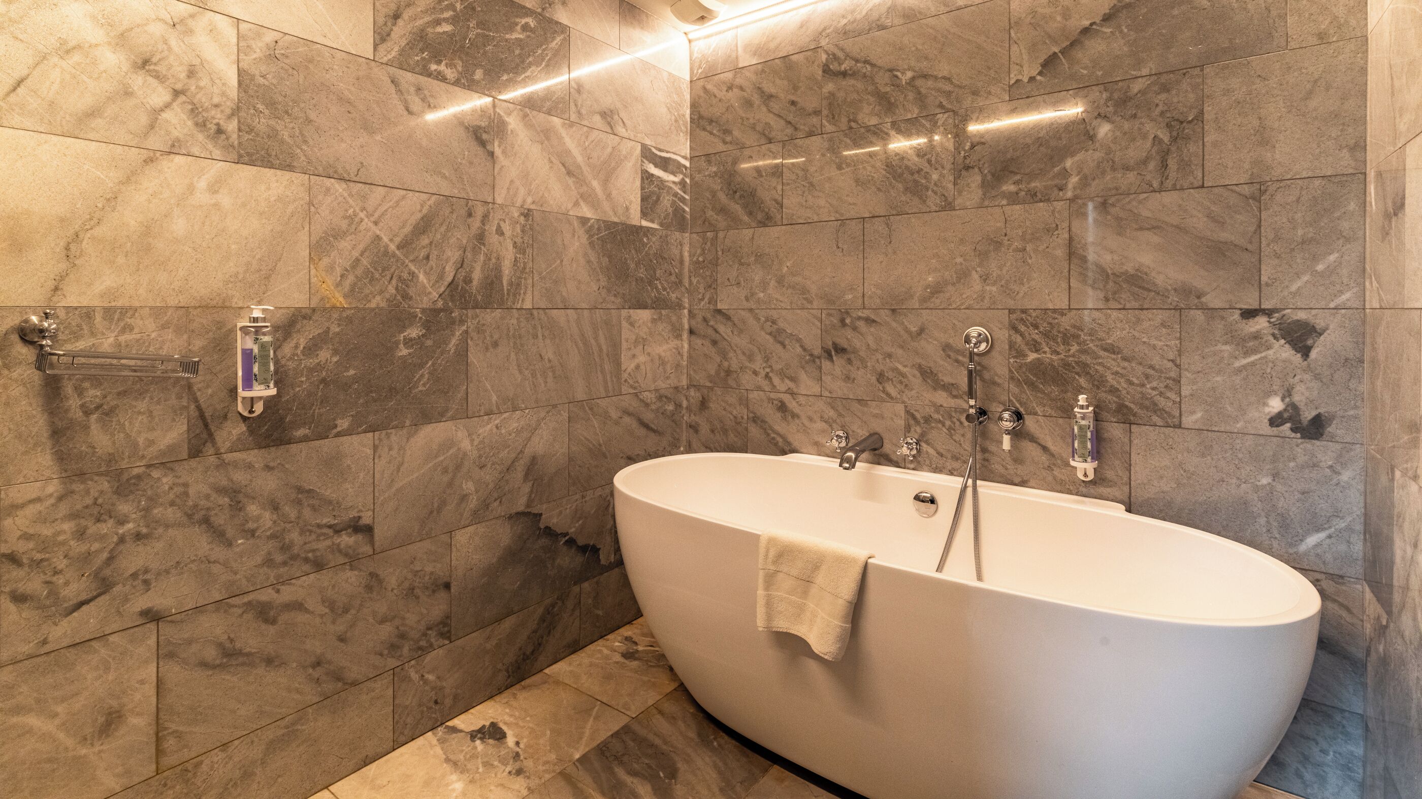 Un baño moderno con una bañera independiente, paredes de mármol y un cabezal de ducha manual.