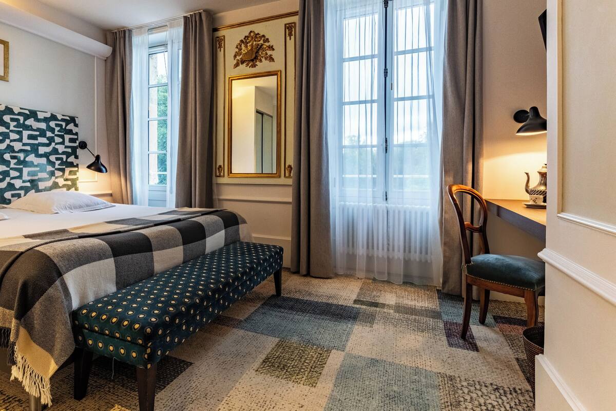 A hotel room with a bed, a bench, a chair, a large mirror, and a window with curtains.