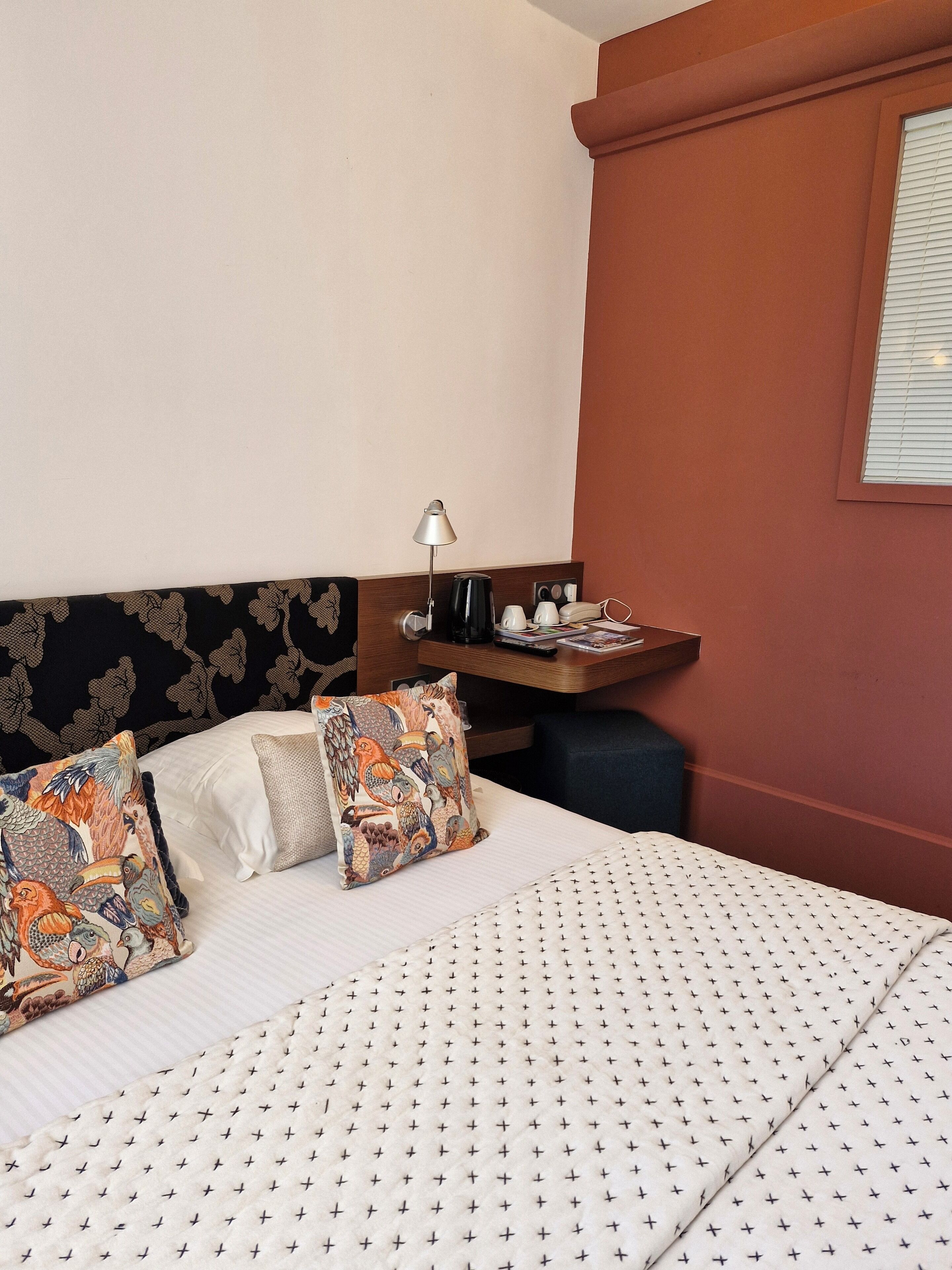 Ett hotellrum med en säng, kuddar, ett sängbord med en lampa, en telefon och ett fönster med persienner.