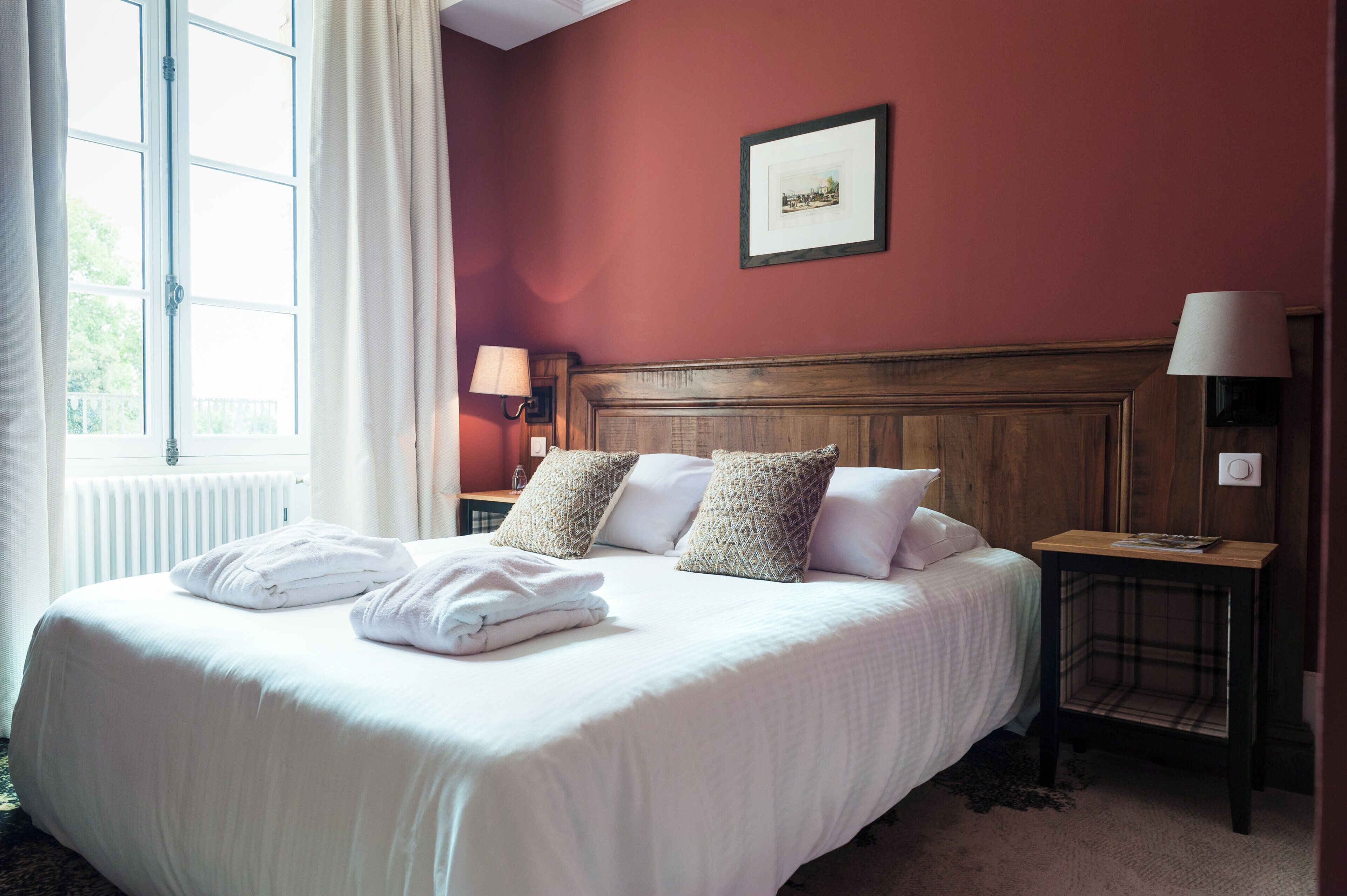 A hotel room with a large bed, a wooden headboard, a nightstand with a lamp, and a window with curtains.