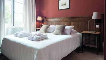 A hotel room with a large bed, a wooden headboard, a nightstand with a lamp, and a window with curtains.