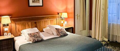 A hotel room with a large bed, two bedside tables with lamps, a wooden headboard, a framed picture on the wall, a door, and a window with curtains.