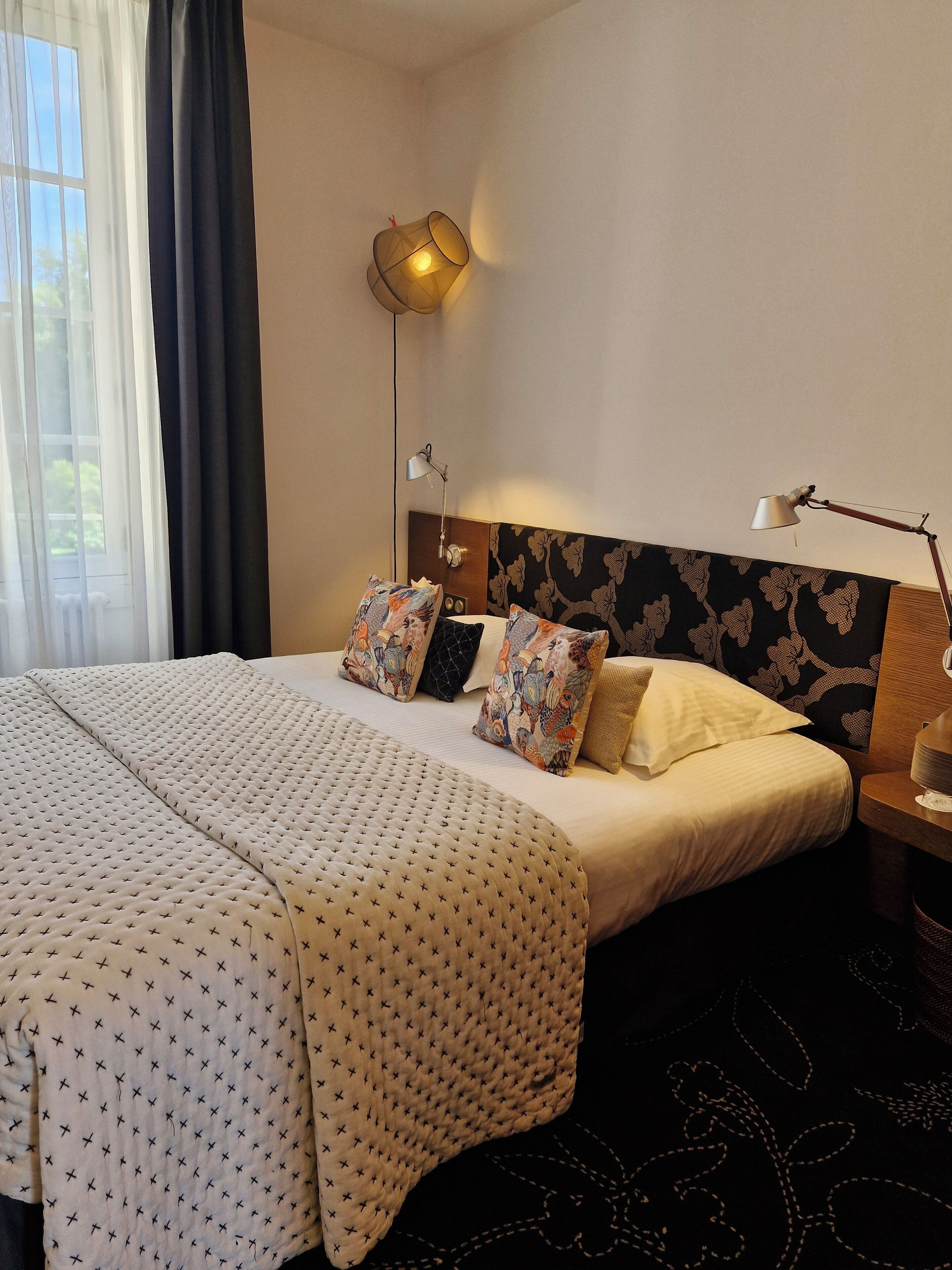 A hotel room with a large bed, a headboard with a floral pattern, two bedside lamps, and a window with curtains.