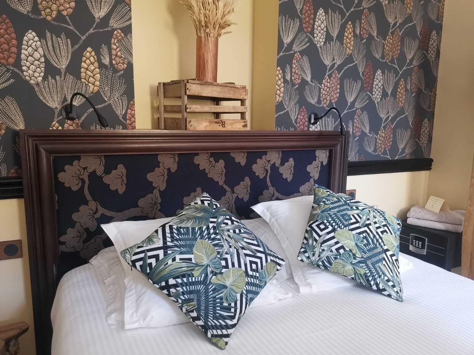 A neatly made bed with patterned pillows, a wooden headboard, and a wall with leaf-patterned wallpaper.