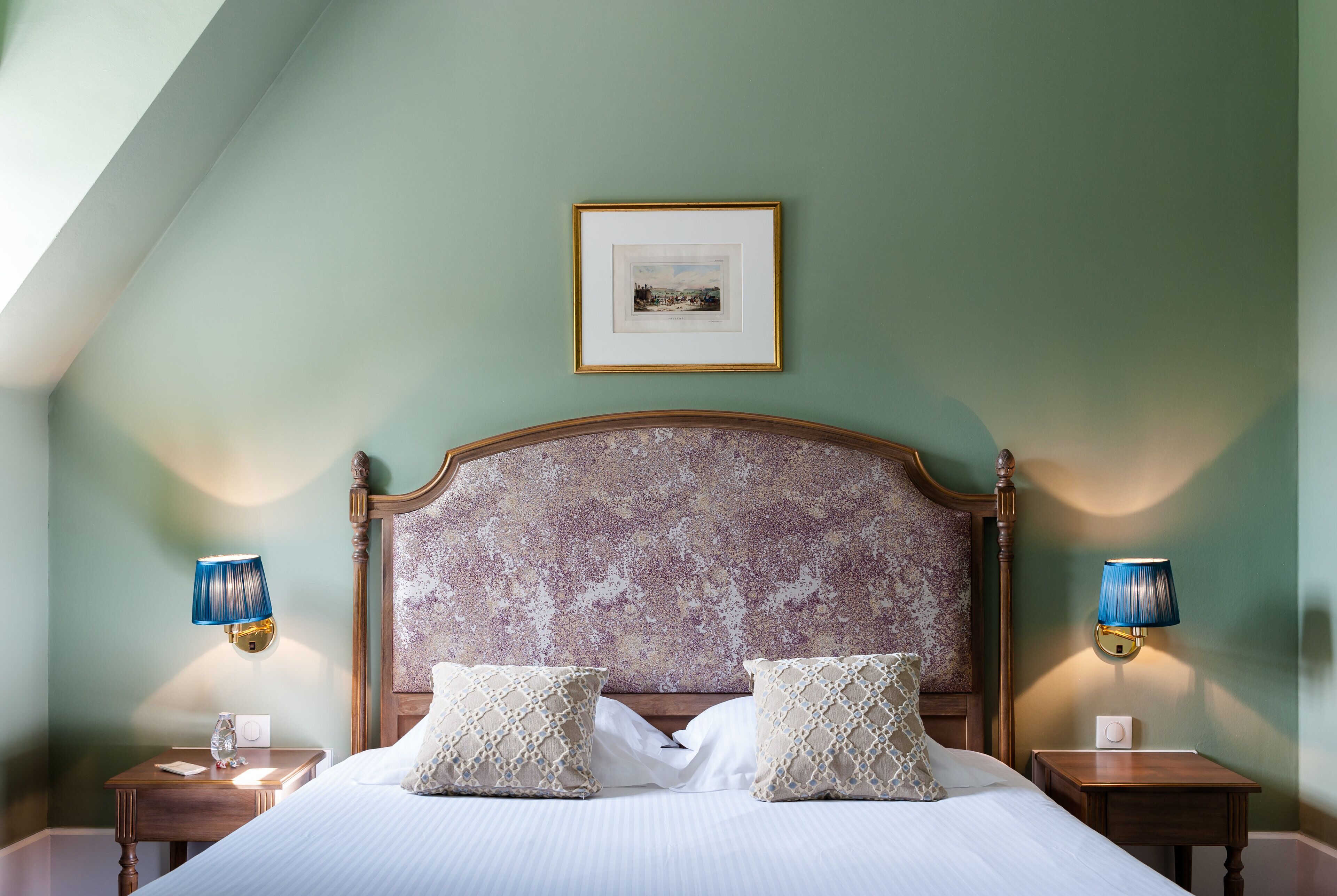 A neatly made bed with two pillows, a framed picture above it, and two bedside lamps on wooden nightstands.