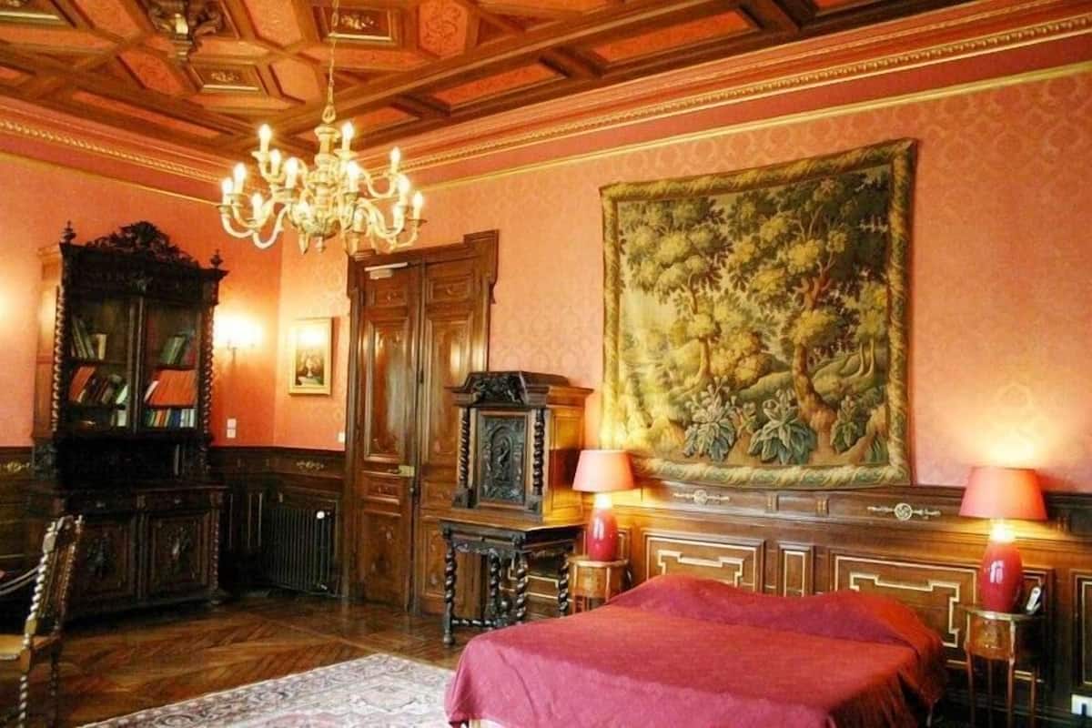 Una habitación con una cama grande, muebles de madera ornamentados, un candelabro y un cuadro grande con motivos florales en la pared.