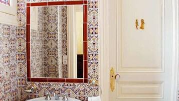 Um banheiro com pia branca, azulejos decorados e estampados, espelho com moldura vermelha e porta branca com puxador dourado.