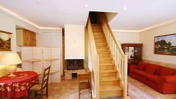 Una sala de estar con escalera de madera, un sofá rojo, una mesa de comedor con mantel rojo, una chimenea y un cuadro en la pared.