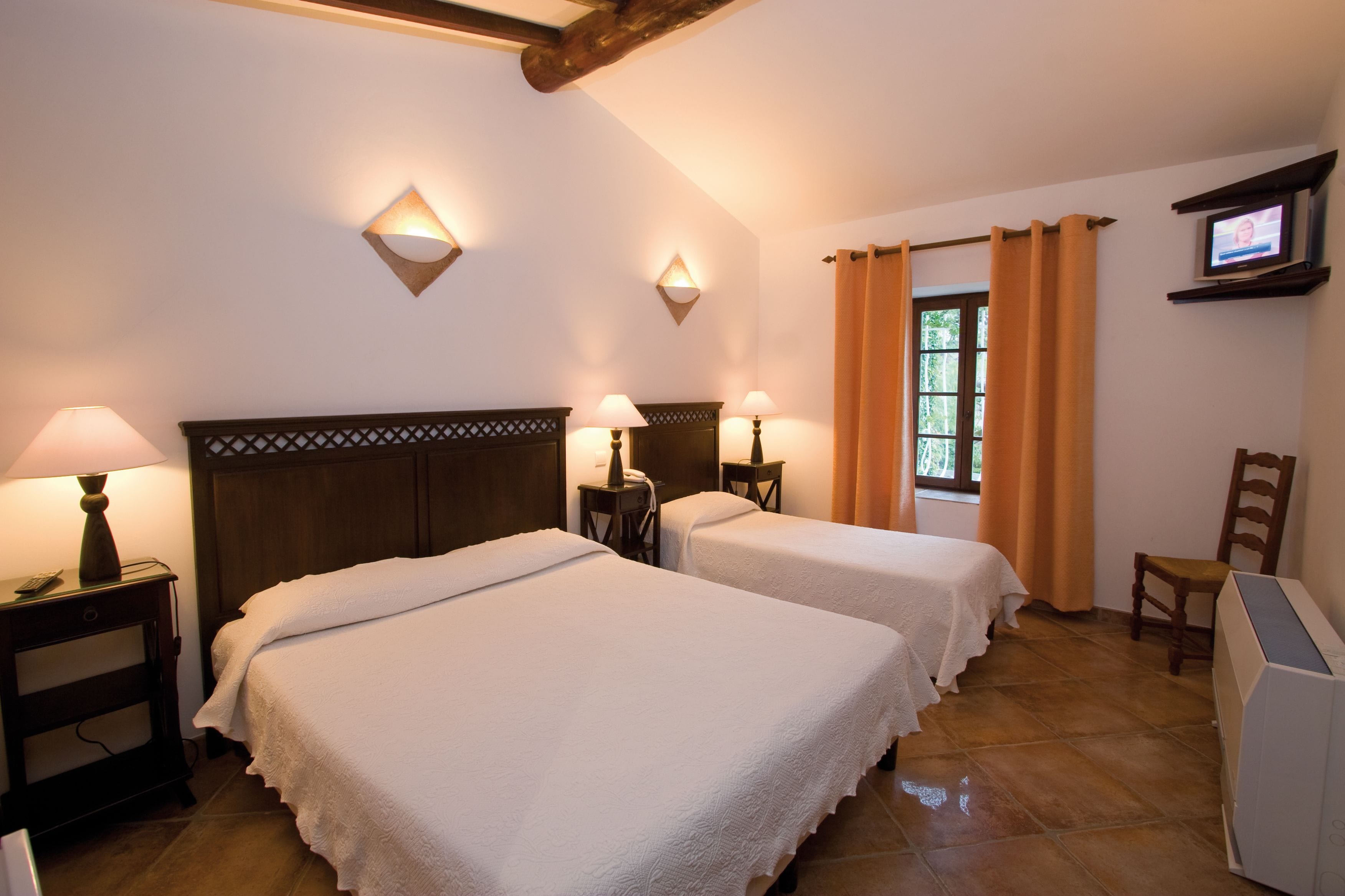 Une chambre d’hôtel avec deux lits, des tables de chevet avec des lampes, un téléviseur posé sur une étagère et une fenêtre avec des rideaux.