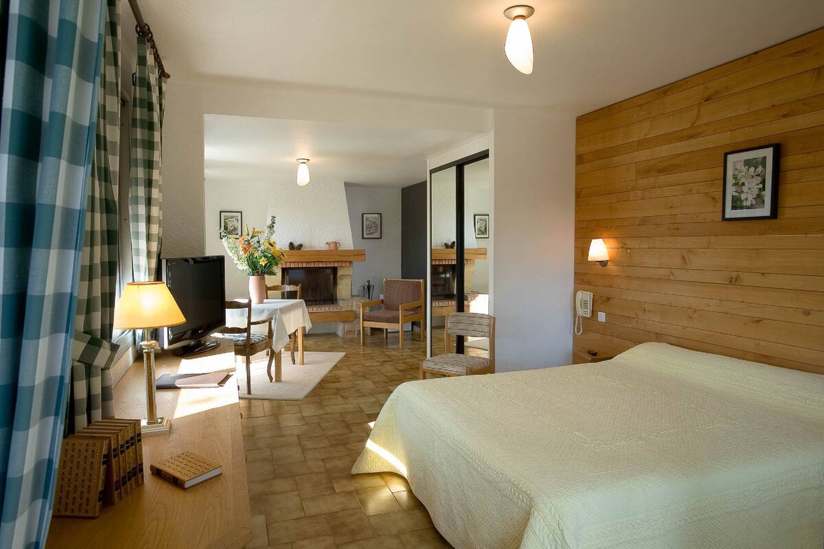 Quarto de hotel com cama, escrivaninha com luminária, televisão e área de refeições com cadeiras e mesa.