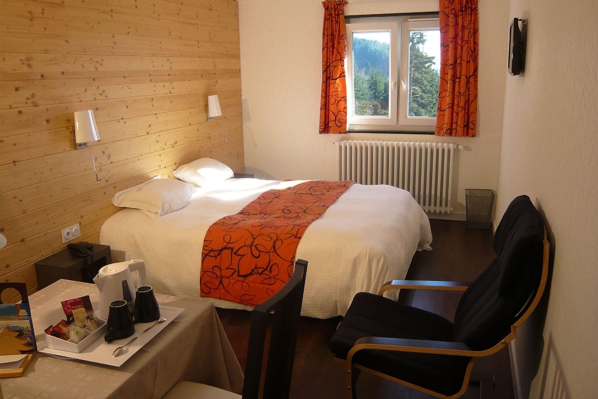 Quarto de hotel com cabeceira de madeira, cama com roupa de cama branca e manta laranja, uma cadeira, mesa com cafeteira e bandeja de lanches, e janela com cortinas laranjas.