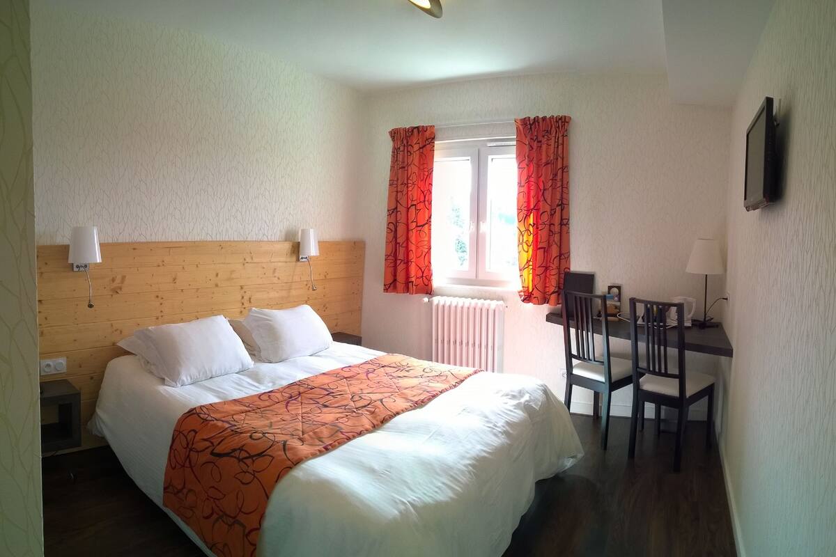 Quarto de hotel com cabeceira de madeira, cama com roupa de cama branca e laranja, escrivaninha com cadeira e janela com cortinas.