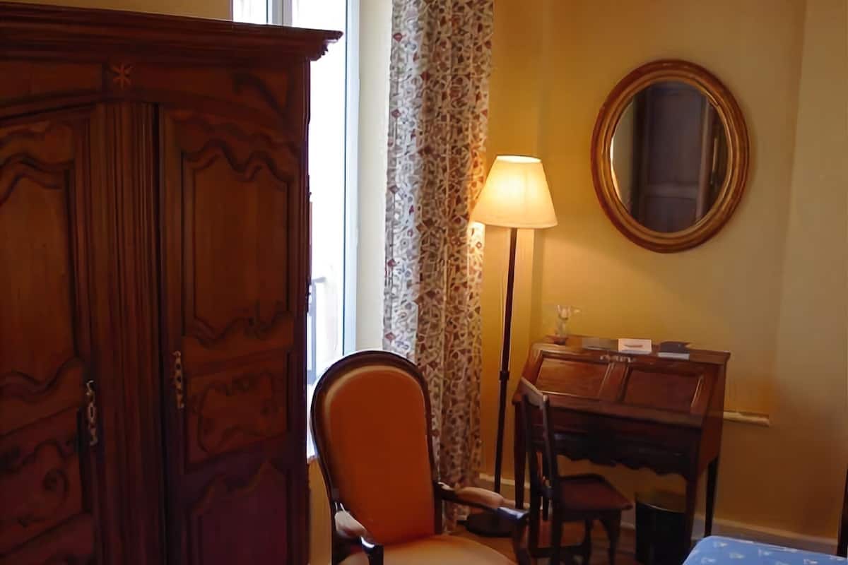 A room with a wooden wardrobe, a chair, a desk, a bed with a patterned bedspread, a lamp, and a mirror on the wall.