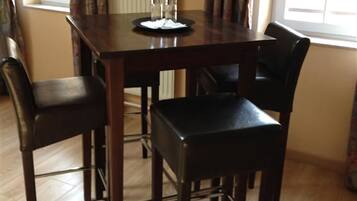 A dining area with a dark wooden table and four chairs. There are two small glasses on the table. The room has a window with curtains and a radiator on the wall.