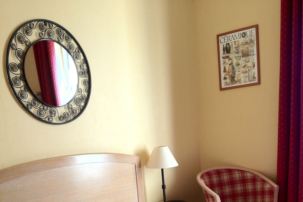 A neatly made bed with a plaid comforter, a bedside lamp, a chair, a wall-mounted mirror, and a ceramic artwork.