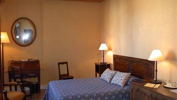 A bedroom with a wooden headboard, a bed with patterned bedding, a desk with a chair, a mirror, and two bedside lamps.