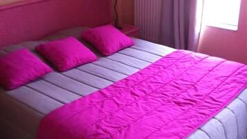 Una cama con una colcha y almohadas rosadas, una lámpara de noche y una ventana con cortinas.