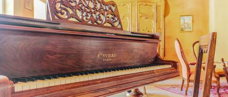 A grand piano with a wooden finish and the brand name 'Caveau Paris' visible on its side, placed in a room with a tiled floor and a wooden chair in the background.