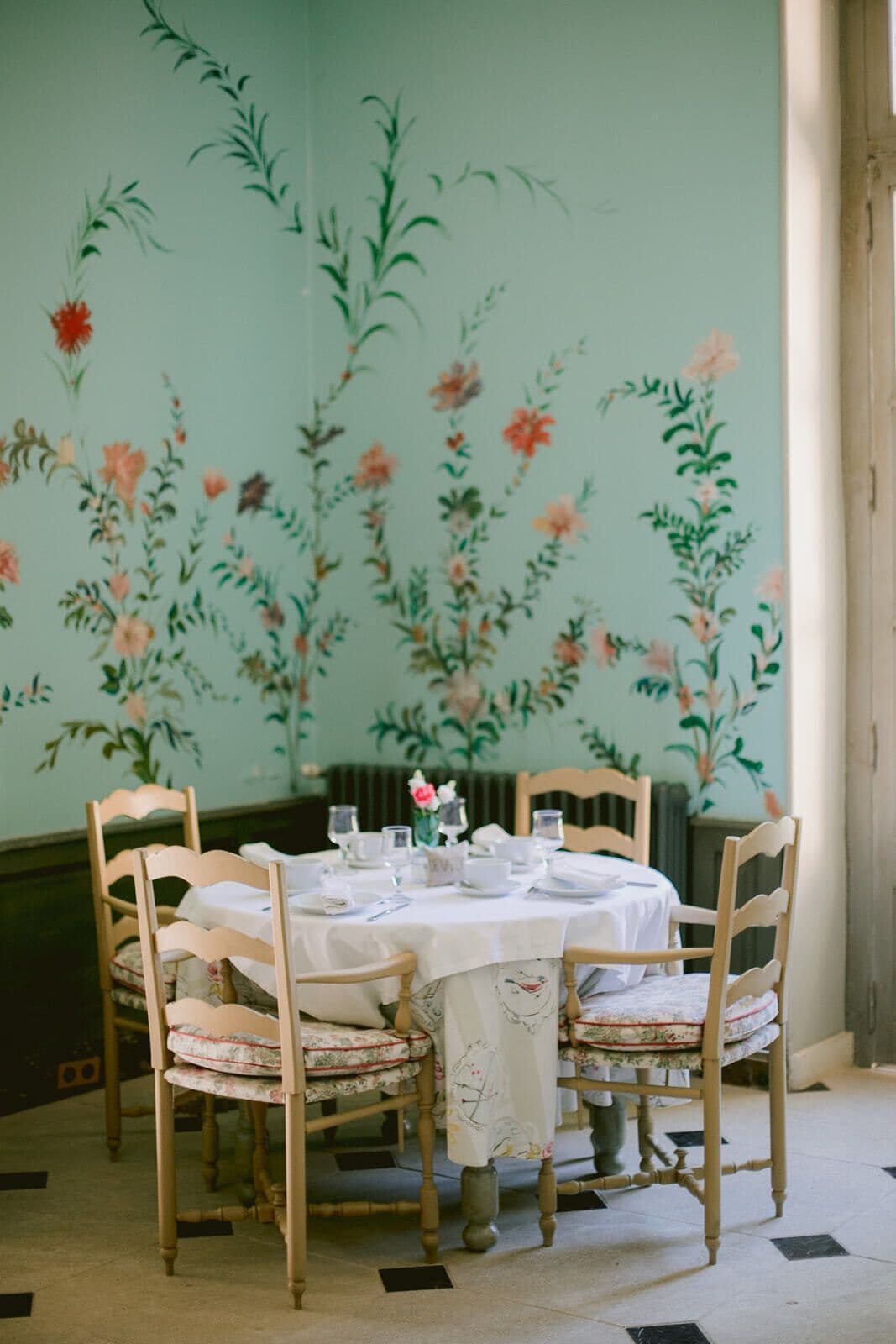 A dining table set for six with floral wallpaper, wooden chairs, and a white tablecloth.