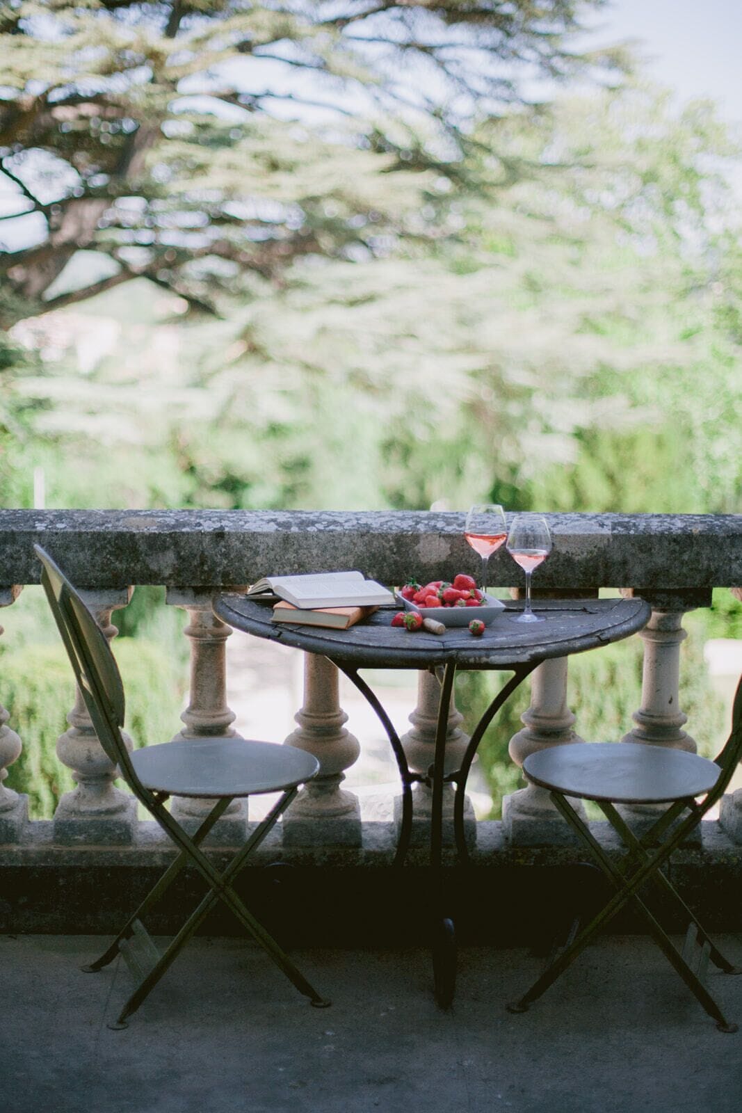 A round table with a book, a bowl of strawberries, and a wine glass on a balcony with stone balustrades and chairs.