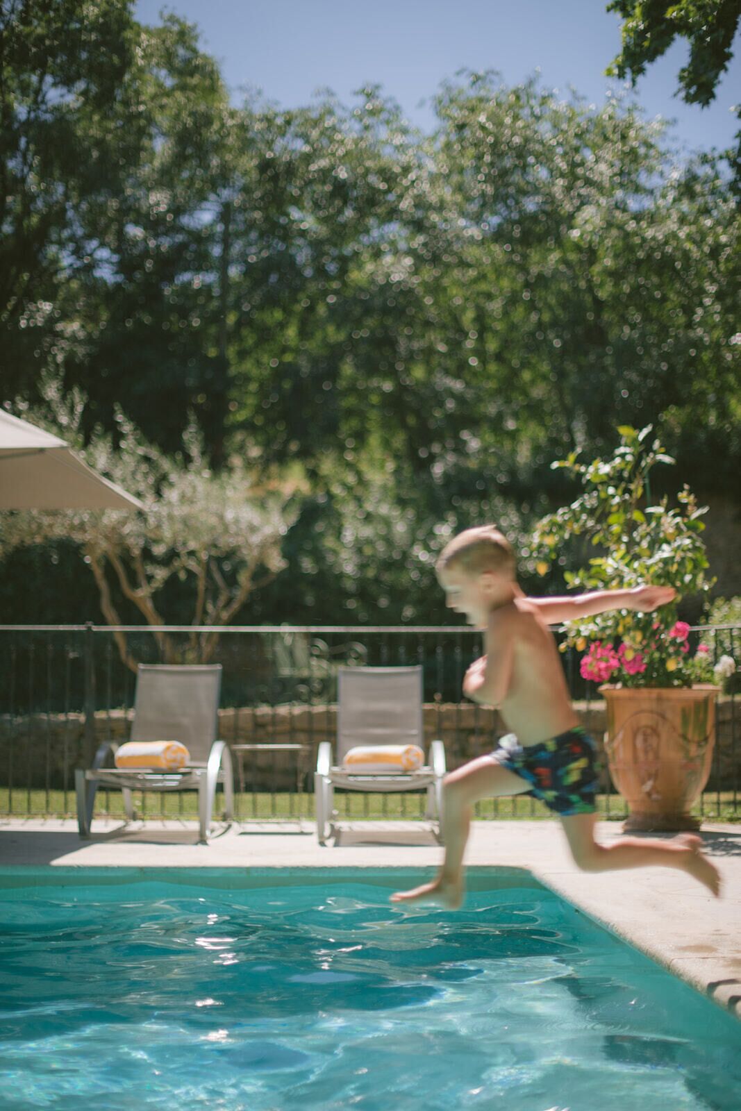 Uma piscina com uma pessoa pulando. Cadeiras de descanso e um vaso de planta ao fundo.