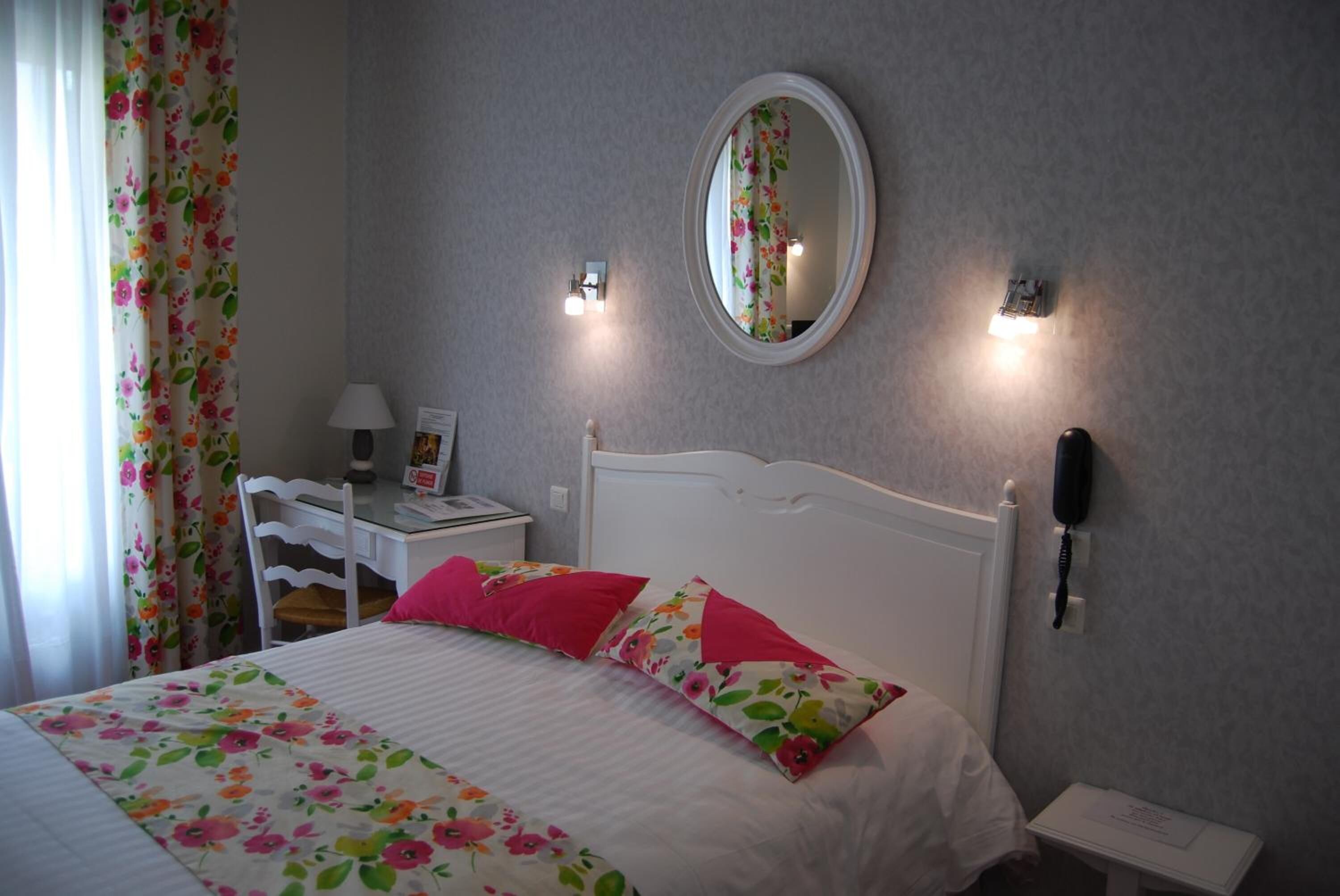 Een slaapkamer met een bed, een bureau, een stoel, een spiegel, een telefoon en een raam met gordijnen.