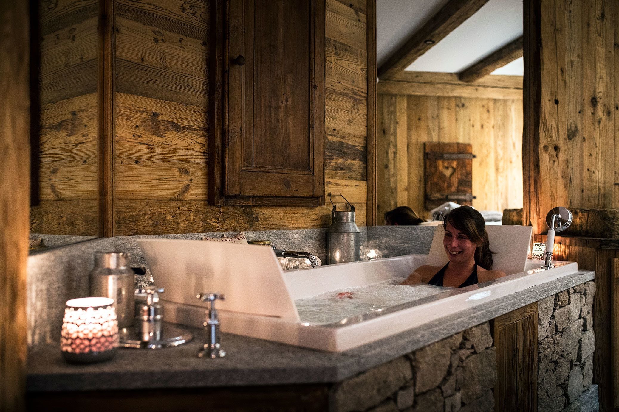 Interior de una cabaña de madera con pared de piedra, una bañera y una vela en la encimera.