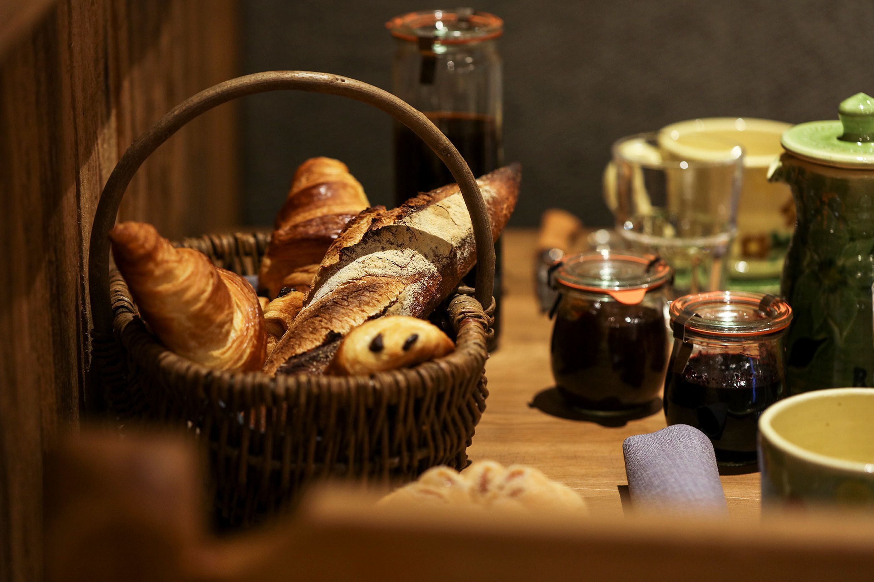 Un panier en osier rempli de différents pains et pâtisseries, une théière et des pots de condiments, le tout posé sur une table en bois.