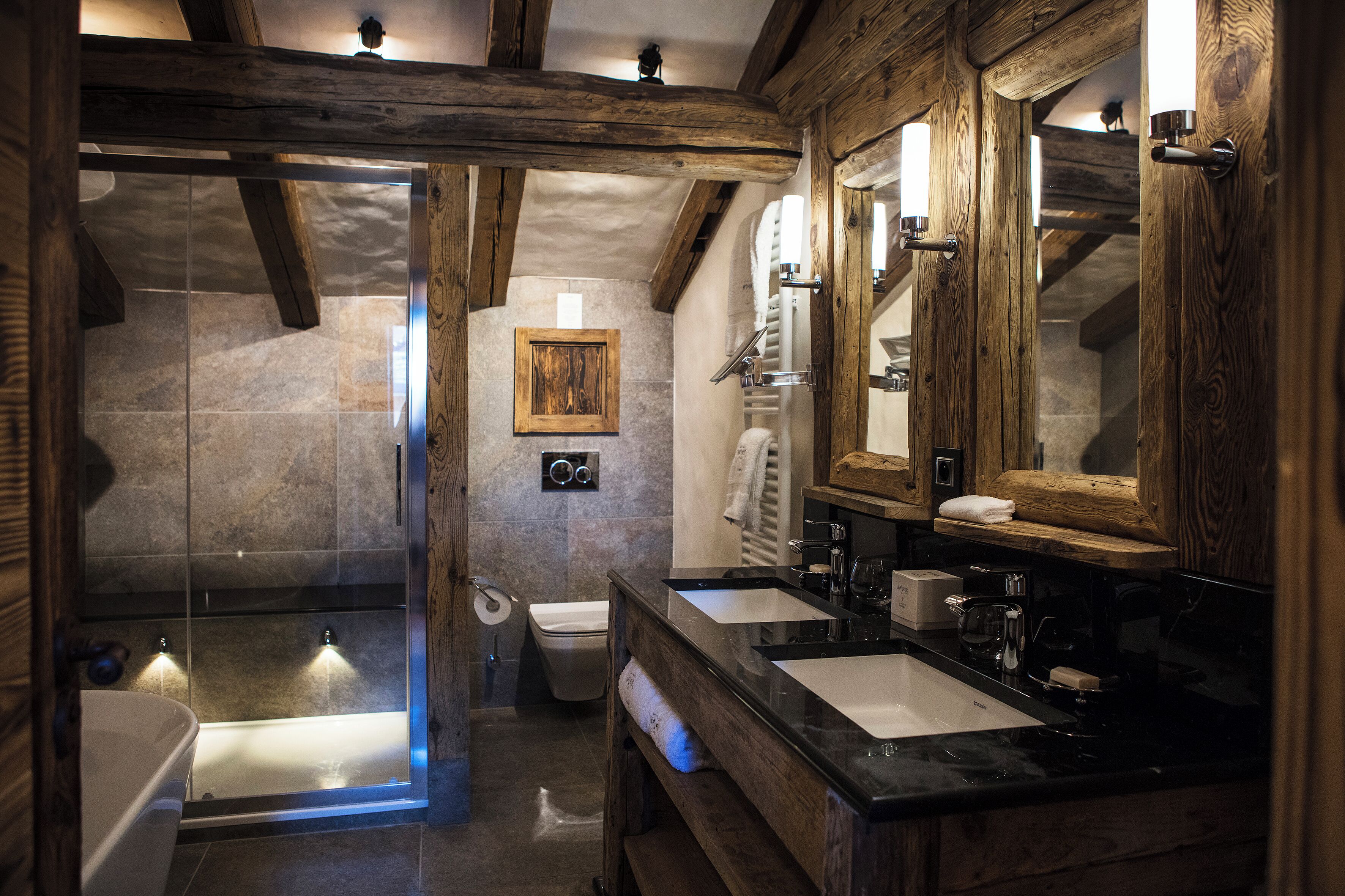 Een badkamer met een douche in een glazen cabine, twee wastafels, houten balken, een grote spiegel en een handdoekhouder.