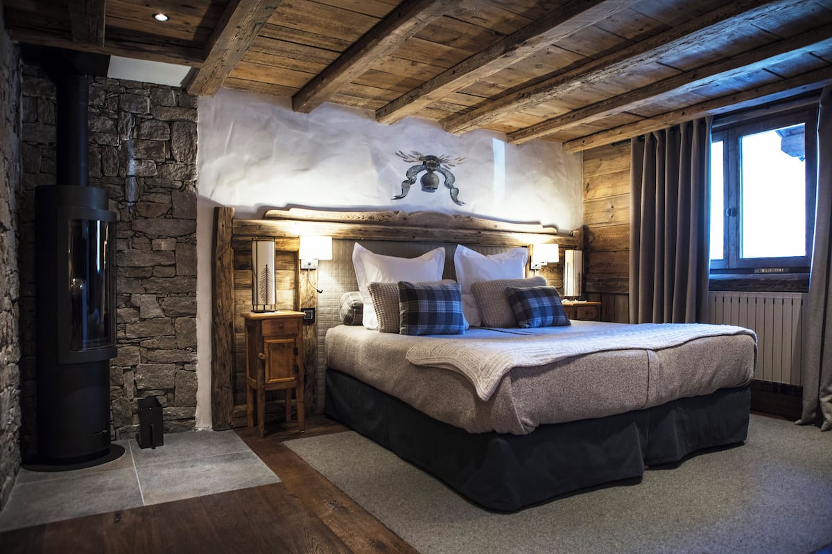 Une chambre confortable avec un mur en pierre, un lit doté d’une tête de lit en bois, une cheminée et une fenêtre avec des rideaux.