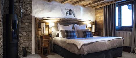 A cozy bedroom with a stone wall, a bed with a wooden headboard, a fireplace, and a window with curtains.