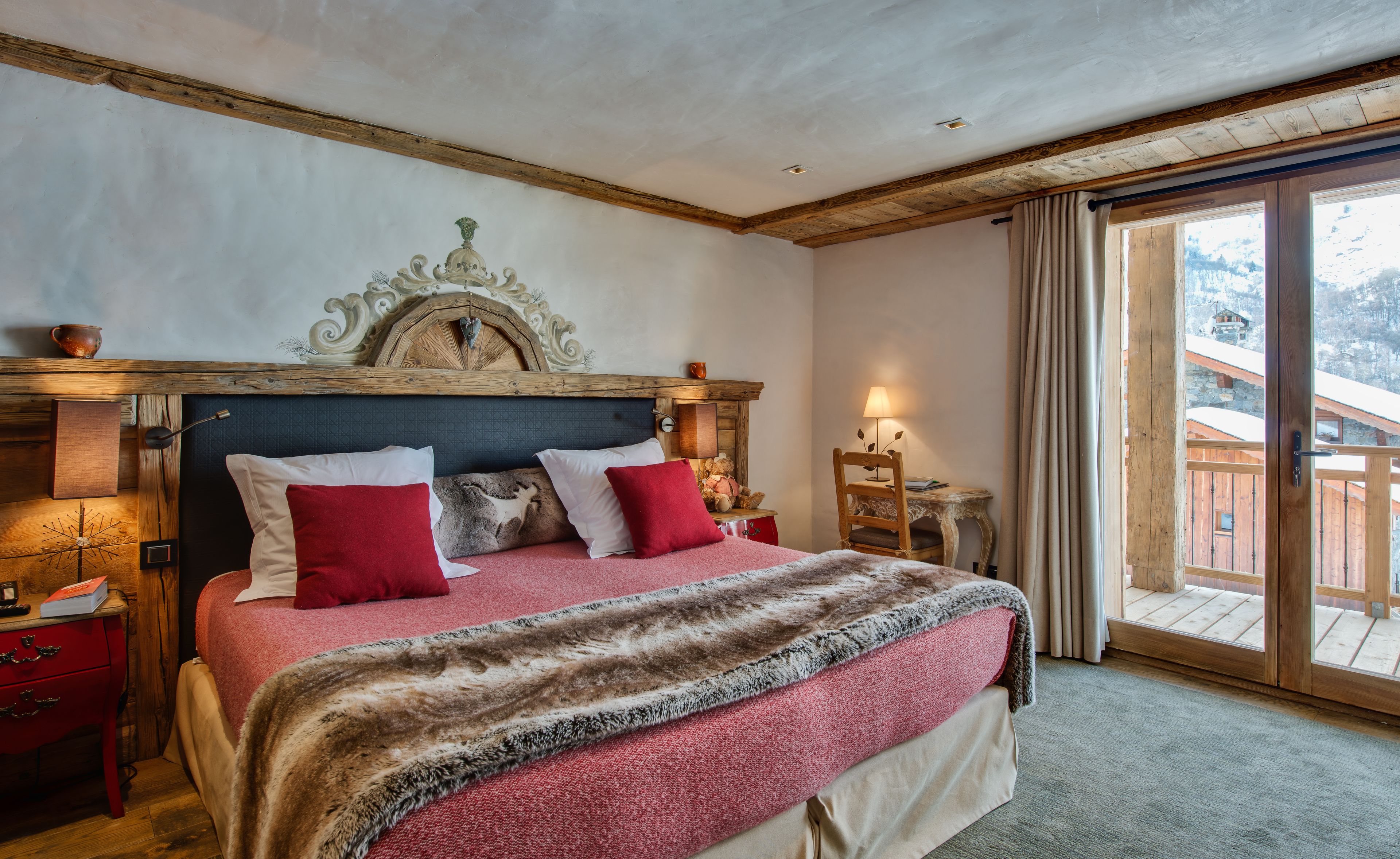 Camera da letto con testiera in legno, un letto grande con lenzuola bianche e rosse, un comodino con una lampada, e vista su una montagna innevata attraverso una porta scorrevole in vetro.