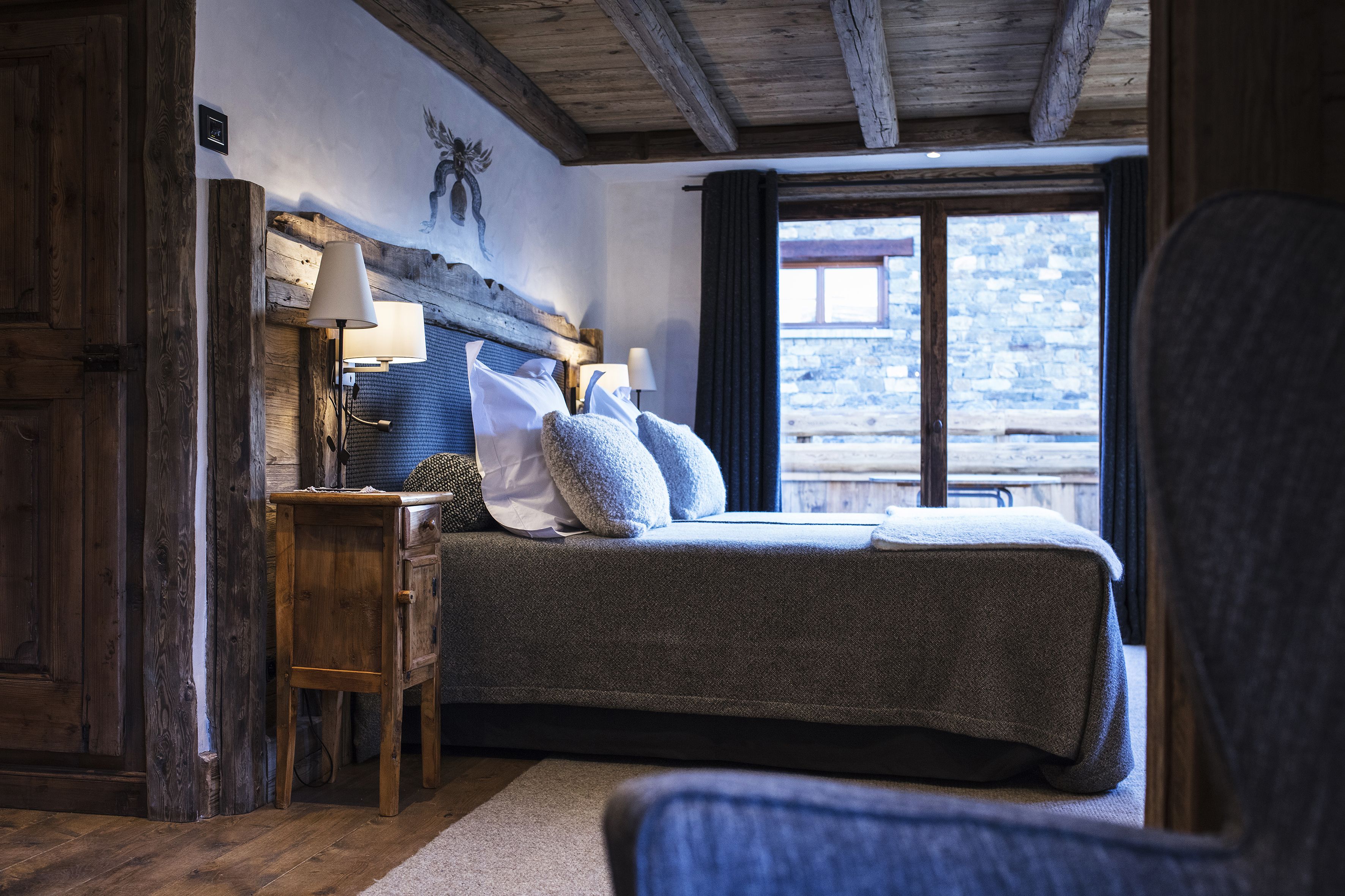 A bedroom with a bed, bedside tables, a lamp, and a view of a stone building through a window.