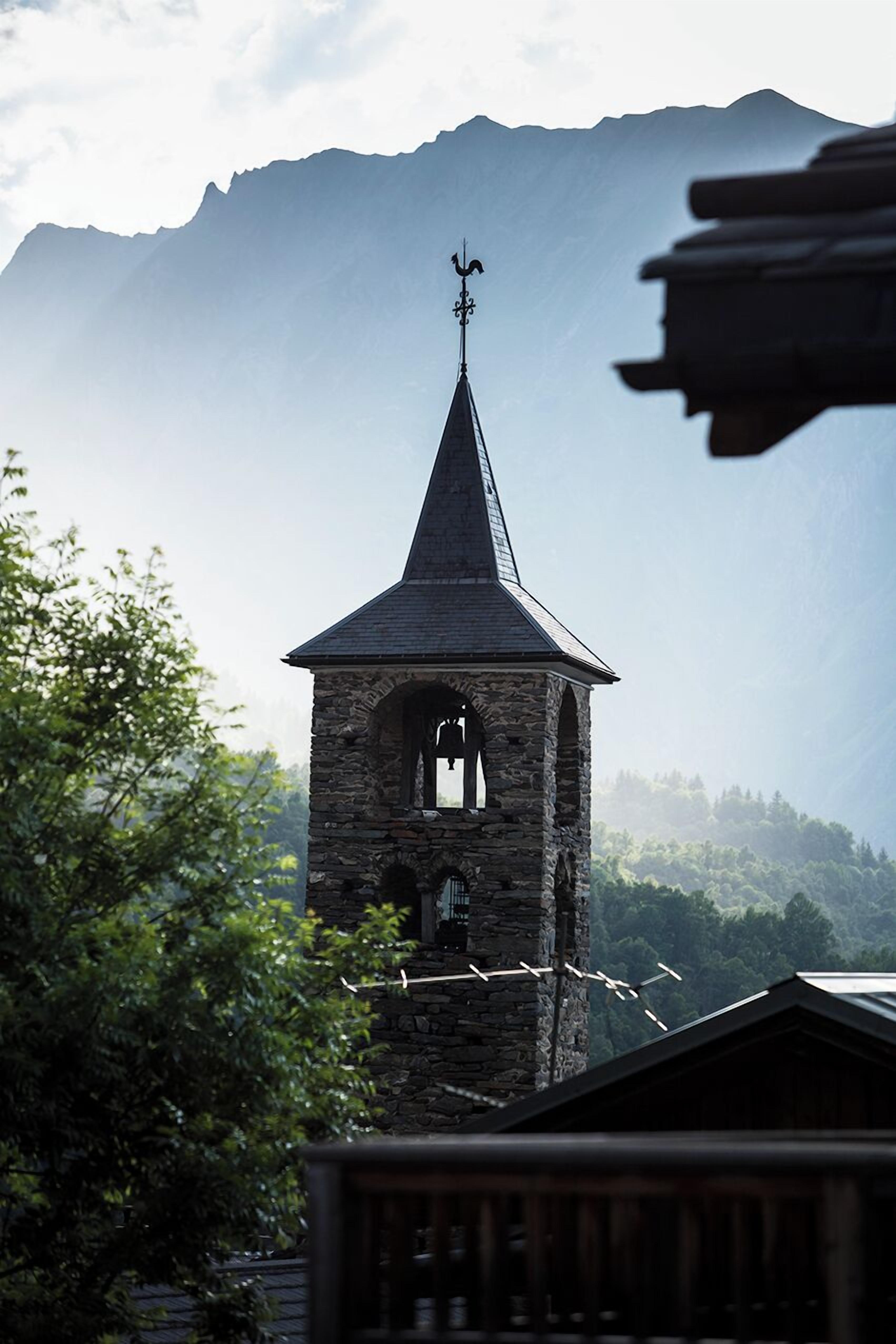 A stone bell tower with a pointed roof and a weather vane, set against a backdrop of mountains and trees.