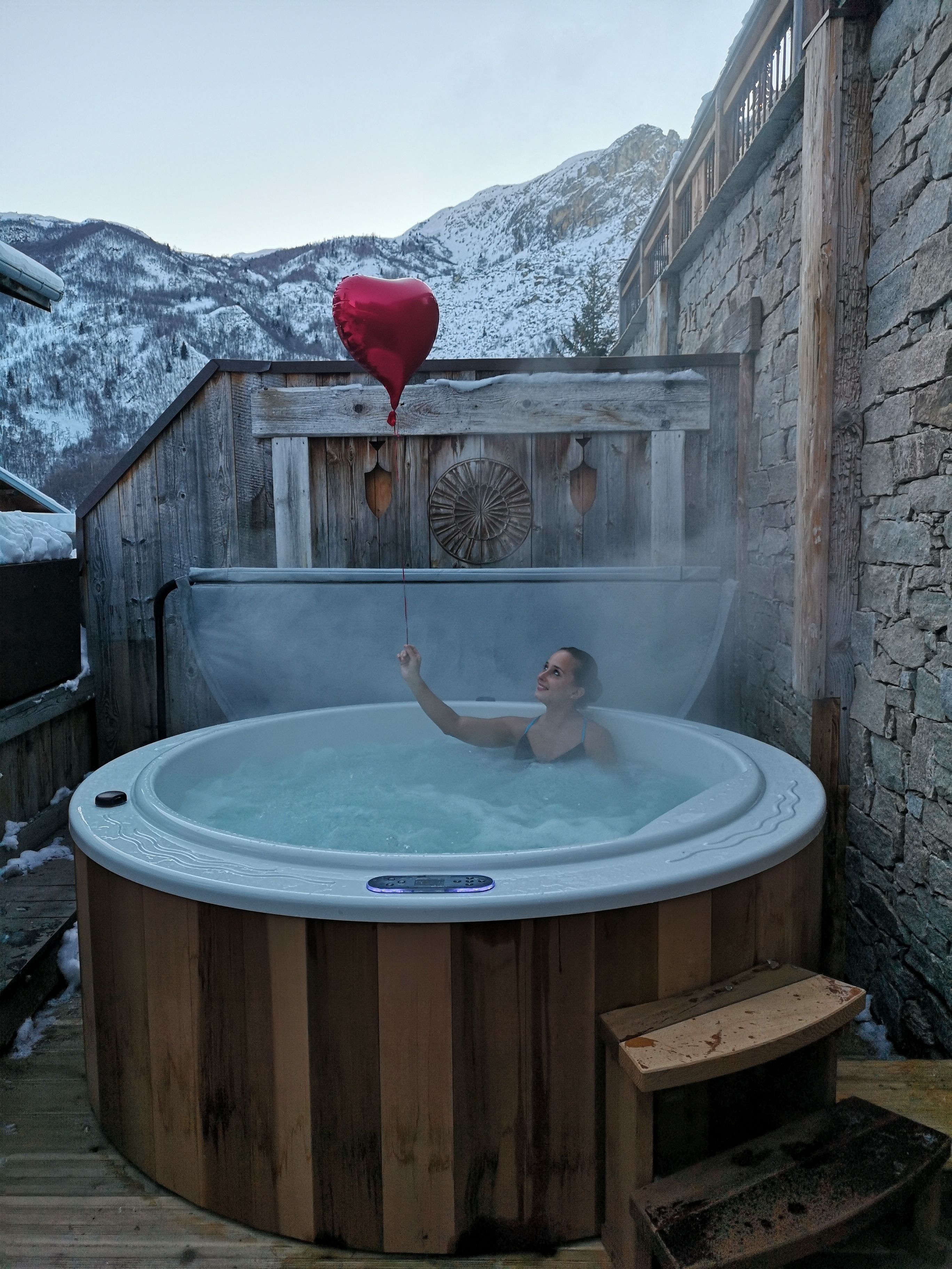 Une personne dans un bain à remous en bois, tenant un ballon rouge en forme de cœur, entourée d’un mur de pierres et avec des montagnes enneigées en arrière-plan.
