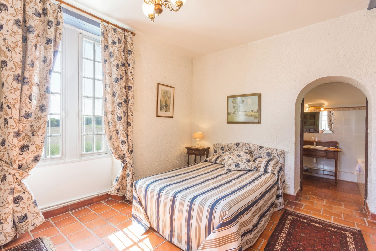 Un dormitorio con cama, cortinas, una mesita de noche, lámpara, un cuadro y un arco que da a un baño.